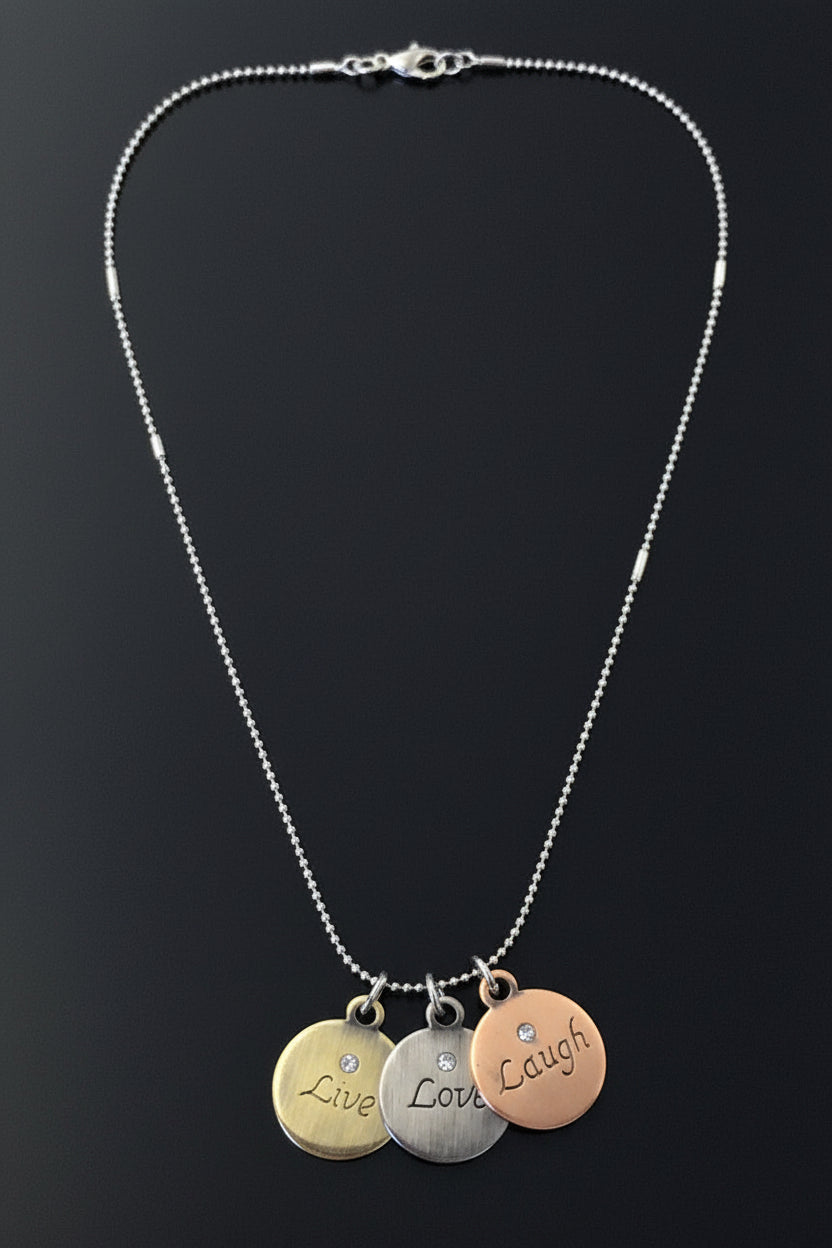 A silver ball-chain necklace with three round engraved charms reading 'Live', 'Love', and 'Laugh', displayed on a black background with a brown label at the bottom.
